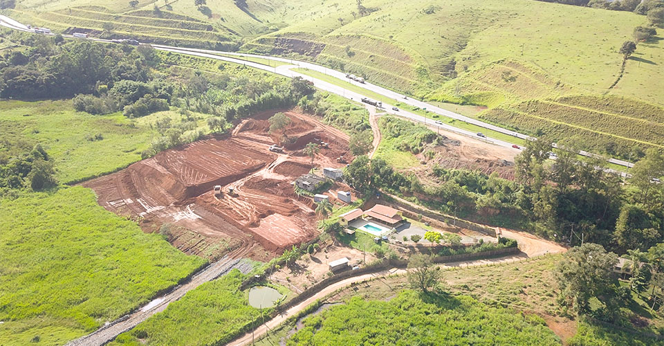 Imagem aérea do início das obras do centro logístico em Camanducaia/MG, com terraplenagem em andamento às margens da rodovia.