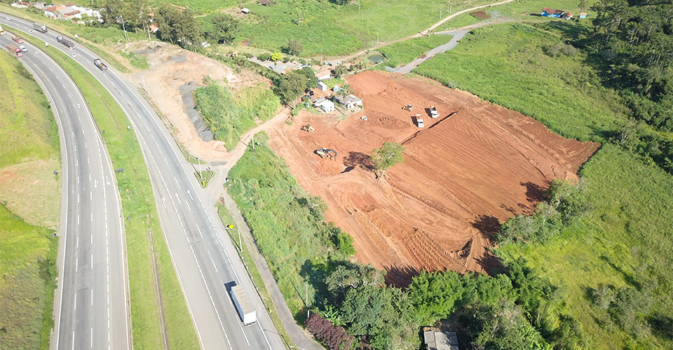 Imagem aérea do início das obras do centro logístico em Camanducaia/MG, com terraplenagem em andamento às margens da rodovia.