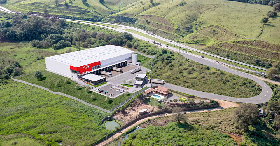 Vista aérea em perspectiva do centro logístico da IMA em Camanducaia/MG, com galpão industrial concluído às margens da rodovia.